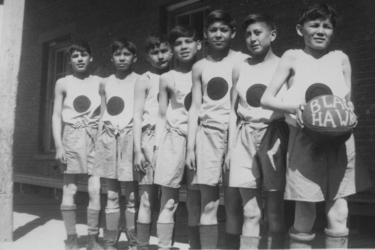 Black and white photo of Indigenous boys lined up in basketball uniforms. The boy closest to the camera holds a basketball.