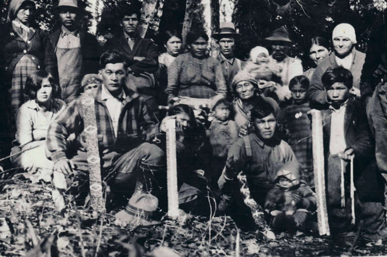 Black and white photo of numerous members of an Algonquin nation seated among trees.