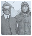 Black and white photo of two men in aviation gear standing against a bi-plane.