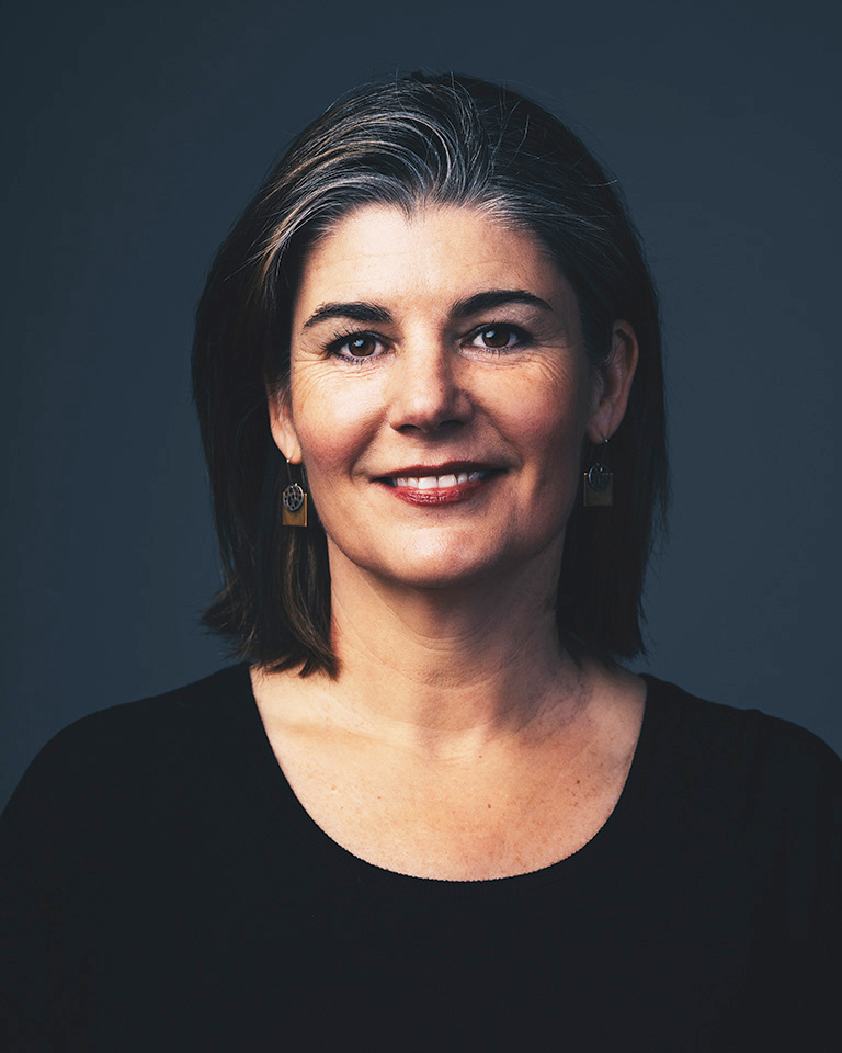 A portrait of a white woman with dark hair. She is in front of a deep blue background wearing a black shirt and earrings.