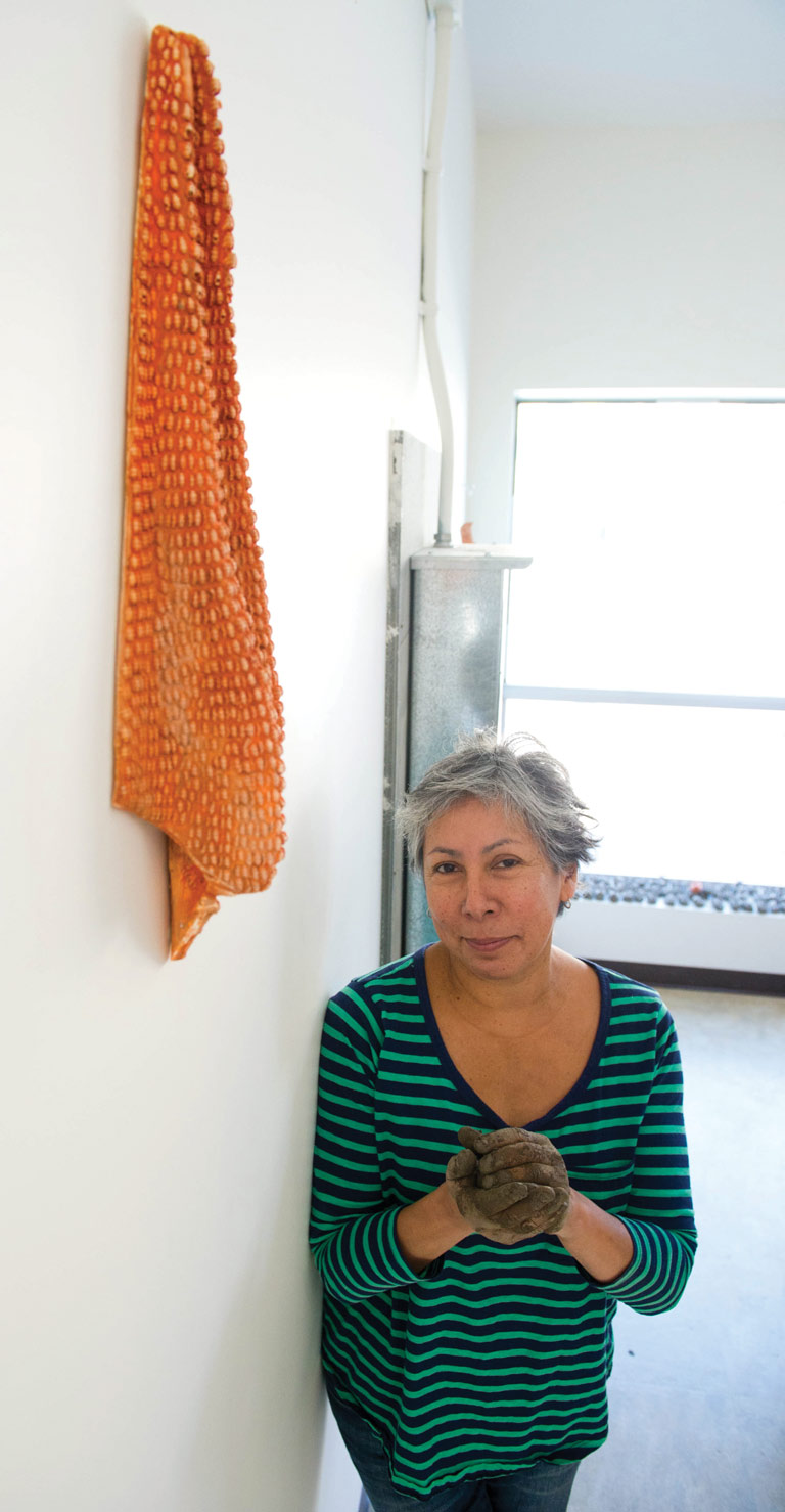 Artist Rebecca Belmore fashions one of thousands of clay "beads" used in her piece Trace. She stands beside a maquette of the artwork, which takes the shape of a large blanket on one of the museum's walls.