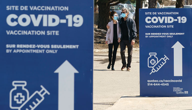 Blue signs lined up along the street read: "Covid-19 Vaccination Site" in French and English. A couple wearing masks walks in-between the signs.
