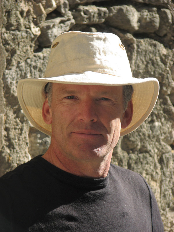 Headshot of Ian Roberts wearing a white hat, black tshirt, in front of a rock wall. 